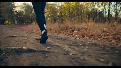 Close-up-a-male-athlete-in-a-black-sports-uniform-and-black-sneakers-runs-along-an-earthen-path-among-fallen-leaves-and-dry-grass-in-an-autumn-forest-on-a-sunny-morning