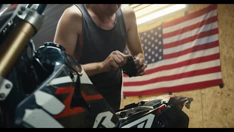 A-male-mechanic-in-a-gray-T-shirt-takes-out-a-metal-part-from-the-back-of-his-motorcycle-in-a-garage---workshop.-Workshop-with-a-US-flag-and-various-tools-for-servicing-equipment