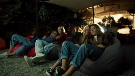 Two-couples-in-love-are-sitting-on-beanbag-chairs-and-watching-a-movie.-A-party-outside-the-city.-Rest-in-the-country-house