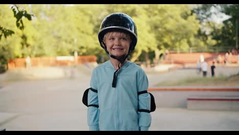 Portrait-of-a-happy-blond-boy-in-a-blue-jacket-in-a-black-helmet-and-elbow-pads,-who-stands-against-the-backdrop-of-a-skatepark-in-the-park