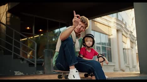 A-dad-with-curly-hair-and-a-Wall-in-blue-jeans-shows-his-little-boy-son-in-a-red-T-shirt-and-a-black-helmet-the-direction-and-they-look-there-together-while-sitting-on-the-floor-near-a-large-building