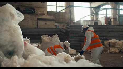 A-man-with-Black-skin-in-a-white-protective-uniform-and-an-orange-vest-together-with-his-colleague,-a-man-with-a-beard-stacks-piles-of-recycled-cellophane-in-one-pile-at-a-waste-processing-and-sorting-plant