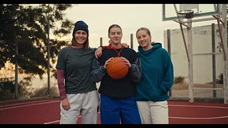 Portrait-of-a-trio-of-happy-girls-basketball-players-blondes-in-hoodies-holding-an-orange-basketball-standing-and-posing-on-a-red-basketball-court-fenced-with-bars-in-the-morning-in-the-summer