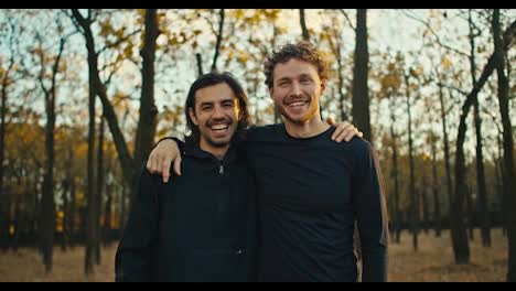 Two-happy-and-cheerful-guys-posing-in-the-autumn-forest-after-their-jog-and-high-fiving-each-other.-Happy-guys-after-a-morning-jog-look-at-the-camera-and-pose-hugging-in-black-sportswear-in-the-autumn-forest