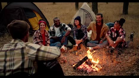 A-group-of-participants-in-a-group-hike-sit-around-a-fire-with-red-coals-and-communicate.-Girls-and-boys-sit-in-a-circle-around-the-fire-enjoying-the-view-and-singing-a-song-to-a-guitar-played-by-a-man-in-a-green-checkered-shirt