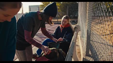 A-trio-of-girls-in-sports-uniforms-put-their-sports-bags-on-a-bench-near-a-lattice-fence-on-a-summer-outdoor-sports-ground-and-prepare-for-basketball-practice-in-the-afternoon