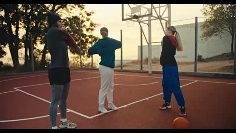 Un-Trío-De-Chicas-Calentando-Antes-De-Su-Práctica-De-Baloncesto-En-La-Cancha-Roja-Cerca-De-Una-Pelota-De-Baloncesto-Naranja-En-Una-Mañana-De-Verano.-Un-Trío-De-Chicas-Preparándose-Para-Jugar-Al-Baloncesto-Y-Calentando-Por-La-Mañana.