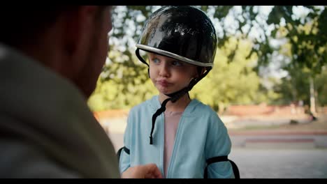 The-father-of-a-little-boy-in-a-blue-T-shirt-with-blue-eyes-helps-him-put-on-a-black-helmet-and-a-blue-jacket.-Little-boy-preparing-to-ride-a-skateboard-outdoor-activity