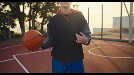 A-close-up-portrait-of-a-blonde-girl-in-a-sports-uniform-who-throws-an-orange-ball-from-hand-to-hand-on-a-red-basketball-summer-street-court-in-the-morning