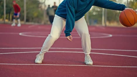 Nahaufnahme-Eines-Blonden-Mädchens-In-Einem-Blauen-Kapuzenpullover-Und-Weißen-Hosen,-Das-Im-Sommer-Einen-Orangefarbenen-Basketball-Auf-Dem-Roten-Boden-Eines-Straßenplatzes-Abprallen-Lässt