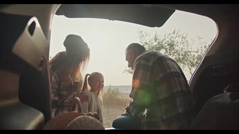 A-happy-man-with-gray-hair-in-a-plaid-shirt-sits-in-the-open-trunk-of-a-car-and-communicates-with-his-little-daughter-and-his-wife,-a-brunette-girl-in-a-plaid-shirt,-during-his-vacation-outside-the-city-in-the-summer