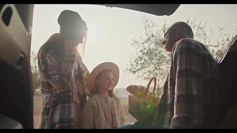 A-happy-family,-middle-aged-man-with-gray-hair-in-a-checkered-shirt-communicates-with-his-little-daughter,-a-blonde-girl,-and-with-his-wife,-a-girl-with-brown-hair-in-a-checkered-shirt.-Sitting-in-the-open-trunk-of-a-car-during-his-vacation-and-picnic-outside-the-city