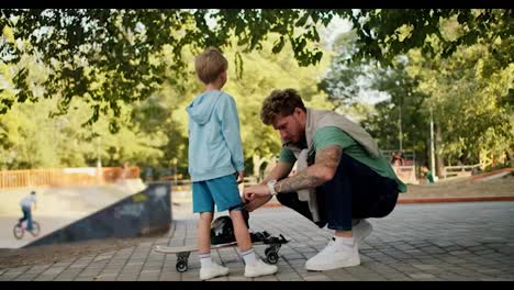 Dad-helps-his-little-son-put-on-protective-clothing-before-riding-a-skateboard-at-the-skatepark-in-the-park.-Father-and-son-lead-an-active-lifestyle