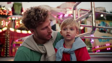 Shooting-in-the-reach,-a-father-with-curly-hair-in-a-Green-T-shirt-communicates-with-his-little-son,-a-blond-man-with-blue-eyes-in-a-red-T-shirt-against-the-backdrop-of-bright-attractions-with-bright-lanterns-and-light-bulbs-in-an-amusement-park.-Dad-and-son-hugging