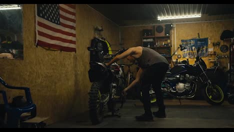 A-male-mechanic-with-a-short-haircut-and-wearing-a-gray-tank-top-puts-his-bike-on-a-jack-in-his-garage-workshop-with-tools-while-repairing-and-working-with-motorcycle-parts