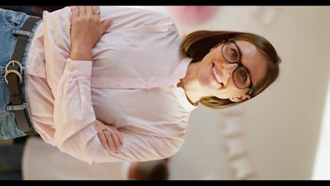 Vertical-video-portrait-of-a-happy-female-teacher-with-a-bob-hairstyle-in-glasses-with-blue-eyes-in-a-pink-shirt-who-smiles-with-her-arms-crossed-on-her-chest-and-looks-at-the-camera-against-the-background-of-a-club-for-preparing-children-for-school