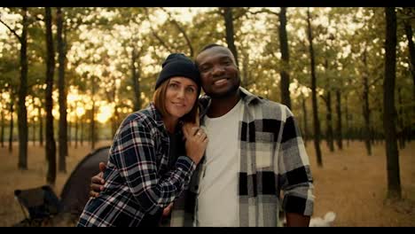 A-happy-couple,-a-guy-with-Black-skin-in-a-checkered-shirt-and-a-girl-with-a-bob-in-a-black-hat,-stand-and-smile-and-look-at-the-camera-during-their-hike-in-a-sunny-populated-summer-forest