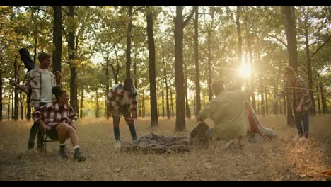 A-happy-team-of-six-people-on-a-hike-arrange-their-things-and-set-up-a-tent-to-spend-the-night-in-a-summer-green-forest-with-withered-grass.-A-break-during-a-group-hike-in-the-summer-forest