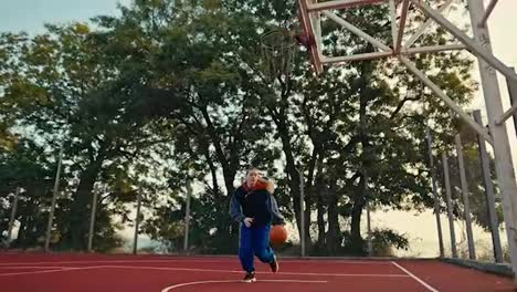 Von-Unten-Aufgenommen-Läuft-Ein-Blondes-Mädchen-In-Einer-Sportuniform-Mit-Einem-Orangefarbenen-Basketballschwert-Und-Wirft-Es-Tagsüber-Auf-Einem-Roten-Straßenplatz-In-Den-Korb