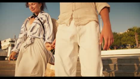 Close-up-shot-of-happy-couple-guy-and-girl-walking-on-modern-beach-holding-hands-in-beachwear-in-summer.-A-guy-in-light-clothes-and-a-brown-haired-girl-go-down-the-stairs-to-the-beach