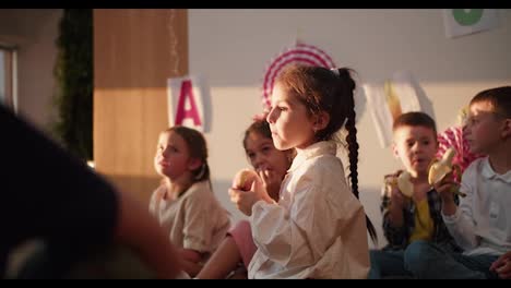 A-little-girl-with-a-braided-hairstyle-in-a-white-shirt-eats-an-apple-at-recess-and-during-lunch-in-a-club-for-preparing-preschool-children-for-school