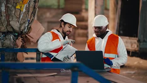 Ein-Brünetter-Mann-Mit-Bart-In-Weißer-Schutzuniform-Und-Orangefarbener-Weste-Spricht-Mit-Seinem-Kollegen,-Einem-Mann-Mit-Schwarzer-Haut,-Der-An-Einem-Laptop-Arbeitet,-Während-Er-In-Einer-Großen-Müllverarbeitungs--Und--sortieranlage-Ist