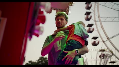 A-dad-with-curly-hair-with-stubble-in-a-green-T-shirt-holds-his-little-blond-son-in-a-red-T-shirt-who-is-shy-and-grabs-his-father's-chest-while-standing-at-the-ticket-office-in-an-amusement-park