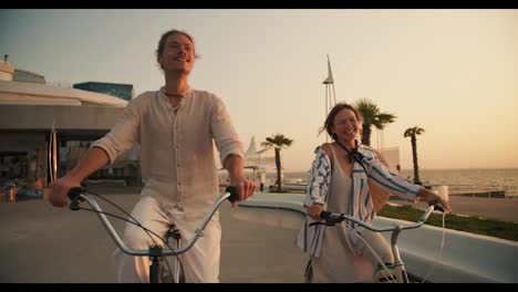 Front-view-of-a-happy-couple,-a-guy-and-a-girl,-ride-bicycles-along-the-beach-on-which-palm-trees-grow-near-the-sea-at-sunrise-in-summer.-Beautiful-modern-beach,-guy-and-girl-riding-bicycles