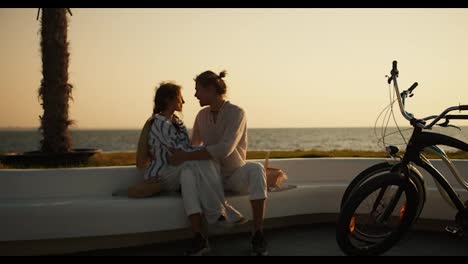 Happy-couple-in-light-clothes-on-a-date-near-the-sea.-A-guy-in-a-light-shirt-and-a-brown-haired-girl-in-a-white-blue-shirt-are-sitting-on-a-white-bench-on-the-beach-near-them-are-their-bicycles-and-the-couple-is-hugging