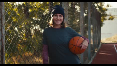 Portrait-of-a-happy-blonde-girl-in-a-black-hat-and-sports-uniform-who-holds-an-orange-basketball-and-leans-on-the-bars-on-a-street-red-court-in-the-morning