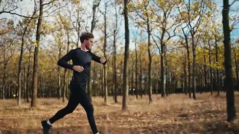 Side-view-of-a-happy-man-with-curly-hair-in-a-black-sports-uniform-running-through-the-morning-autumn-forest-with-fallen-leaves