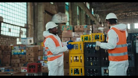 A-man-with-Black-skin-in-a-white-protective-uniform-together-with-his-colleague-a-brunette-man-with-a-beard-in-an-orange-vest-puts-glass-bottles-in-special-containers-and-communicates-while-they-work-at-a-waste-recycling-plant