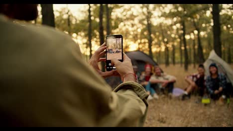 Un-Hombre-Con-Una-Camisa-Verde-Claro-Toma-Fotografías-De-Su-Compañía-Con-La-Que-Fue-De-Excursión-Usando-Una-Cámara-En-Su-Teléfono-En-Un-Bosque-Soleado-De-Verano.-Tomando-Una-Foto-Para-El-Recuerdo-Durante-Una-Parada-Para-Acampar