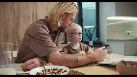 Ein-Blonder-Mann-Mit-Bart-Und-Brille-Nimmt-Einen-Bleistift-Und-Zeigt-Seinem-Kleinen-Sohn,-Was-Er-Bei-Den-Hausaufgaben-Tun-Muss.-Seinem-Albino-Sohn-Gefällt-Das-Nicht.-Bei-Den-Schulvorbereitungen-In-Der-Küche-Einer-Modernen-Wohnung-Kommen-Sie-Jedoch-Zu-Einer-Gemeinsamen-Lösung-Des-Problems.