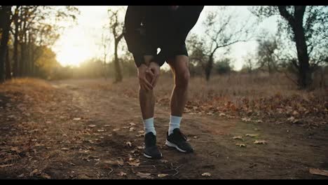 Un-Chico,-Un-Deportista-Con-Uniforme-Deportivo-Negro,-Mientras-Trota-En-El-Bosque-Se-Agarra-La-Rodilla-Y-Se-La-Frota-Porque-Siente-Daño-Y-Lesión.