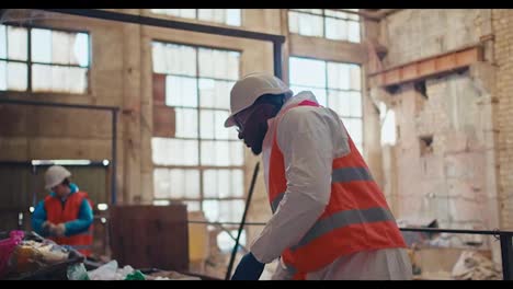 A-man-with-Black-skin-in-a-special-white-protective-uniform-and-an-orange-vest-sorts-plastic-at-an-old-waste-processing-plant.-A-man-with-Black-skin-is-an-employee-of-a-huge-waste-recycling-plant