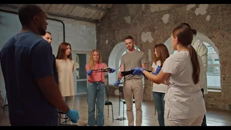 Shooting-behind-the-shoulders:-A-professional-female-doctor-in-a-medical-white-uniform,-together-with-a-Black-man-assistant-in-a-blue-medical-uniform,-conduct-a-medical-training-course,-talking-about-medical-harnesses-and-showing-them-to-the-public-against-the-background-of-white-brick-walls