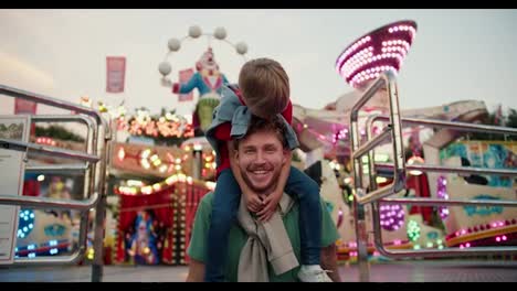 Portrait-of-a-Happy-father-with-curly-hair-in-a-Green-T-shirt-and-his-little-blond-son-in-a-red-T-shirt-against-the-backdrop-of-bright-lights-and-attractions-in-an-amusement-park