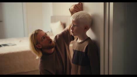 A-blond-car-with-glasses-and-a-beard-measures-the-height-of-his-little-albino-boy-with-white-hair-who-stands-near-the-wall-and-waits-for-his-dad-to-tell-him-his-height