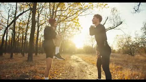 A-man-with-curly-hair-in-a-black-sports-uniform-together-with-his-friend-in-black-shorts-and-a-red-hat-are-doing-sports-and-warming-up-before-their-jog-in-the-autumn-forest-in-the-evening-at-sunset-on-an-earthen-path