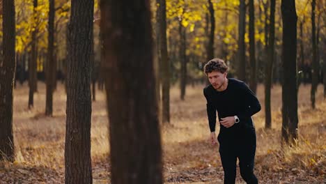 Un-Hombre-Con-Cabello-Rizado-Y-Barba-Con-Uniforme-Deportivo-Negro-Corre-En-El-Bosque-Otoñal-Por-La-Mañana-Durante-Su-Trote-Entre-Los-árboles-En-El-Bosque-Soleado.