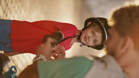 Vertical-video-of-a-dad-with-curly-hair-in-a-Green-T-shirt-helping-his-little-blond-son-with-blue-eyes-in-a-red-T-shirt-put-on-protective-clothing-before-riding-a-skateboard-in-the-park