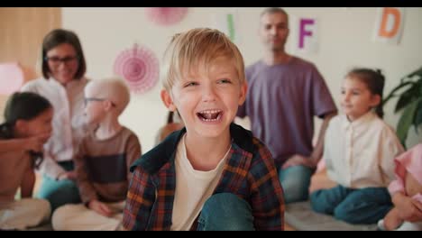 Porträt-Eines-Glücklichen-Und-Fröhlichen-Jungen-Mit-Blauen-Augen-In-Einem-Karierten-Hemd,-Der-Vor-Dem-Hintergrund-Seiner-Ersten-Unterrichtsstunde-Sitzt-Und-Lächelt,-Um-Sich-Zusammen-Mit-Seinen-Lehrern-Und-Einer-Gruppe-Von-Kindern-Auf-Die-Schule-Vorzubereiten