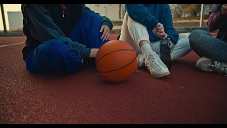 Close-up-three-girls-in-sportswear-are-holding-sports-bottles-and-sitting-on-the-red-floor-near-an-orange-ball-on-a-basketball-street-court-in-the-morning