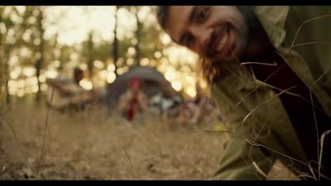 A-happy-brunette-man-in-a-light-green-jacket-sets-up-a-video-camera-and-then-goes-to-the-rest-of-the-group-of-hikers-and-poses-with-them-taking-a-delayed-photo.-Happy-group-photo-of-hiking-participants-in-hiking-clothes-against-the-backdrop-of-tents-along-with-a-guitar-and-a-light-green-sunny-summer