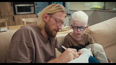 A-blond-man-in-glasses-with-a-beard-shows-his-little-albino-son-in-blue-glasses-how-to-write-letters-in-English-correctly-and-also-how-to-properly-prepare-for-his-first-lesson-at-school.-Happy-blond-man-and-his-little-albino-son-laughing-and-having-fun-while-doing-homework