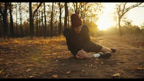 A-man-in-a-black-sports-uniform-with-a-red-cap-makes-a-face-and-holds-his-knee-while-jogging-in-the-forest-he-lies-on-the-ground.-Morning-jogging-safety-rules