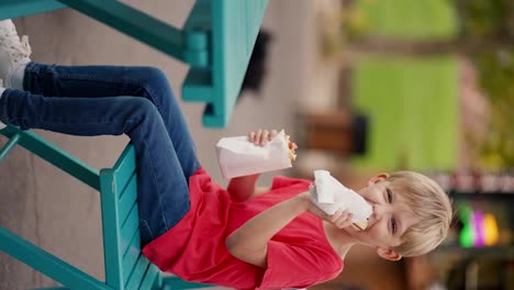 Vertical-video-A-little-blond-boy-with-blue-eyes-in-a-red-T-shirt-eats-hot-dogs-on-a-street-table-near-street-cafes-in-the-park.-Happy-little-boy-has-a-snack-in-the-park