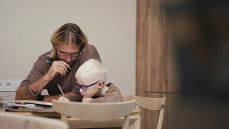 Ein-Blonder-Mann-Mit-Bart-Und-Brille-Hilft-Seinem-Kleinen-Albino-Sohn-Mit-Weißer-Haarfarbe-Und-Blauer-Brille,-Seine-Hausaufgaben-Zu-Machen-Und-Alles-Richtig-In-Mathematik-Zu-Berechnen,-Während-Er-Zu-Hause-In-Einer-Modernen-Küche-Hausaufgaben-Macht