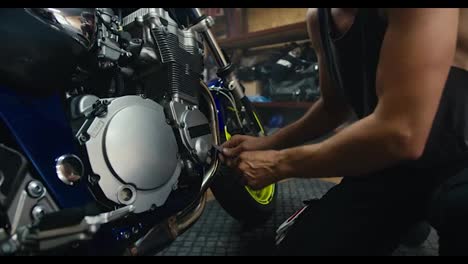 Close-up-shot-of-a-male-mechanic-in-a-gray-T-shirt-using-a-tool-to-repair-his-blue-motorcycle.-A-man-with-strong-arms-with-visible-veins-works-in-a-workshop